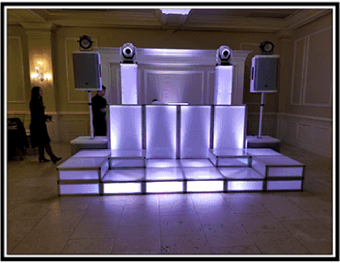 DJ stage with white LED panels and speakers, indoors. Person operating equipment, another standing nearby.