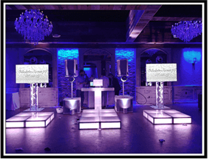 Three illuminated photo booths on platforms in a blue-lit event space with chandeliers and DJs.
