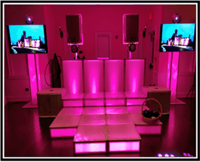A DJ setup lit with pink lights, including speakers, screens, and a stage.
