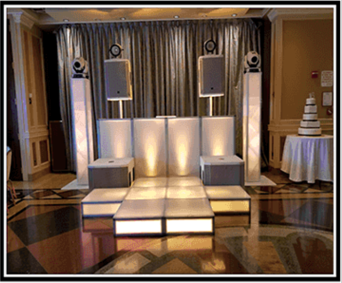 DJ stage setup with illuminated panels and speakers, near a wedding cake table.