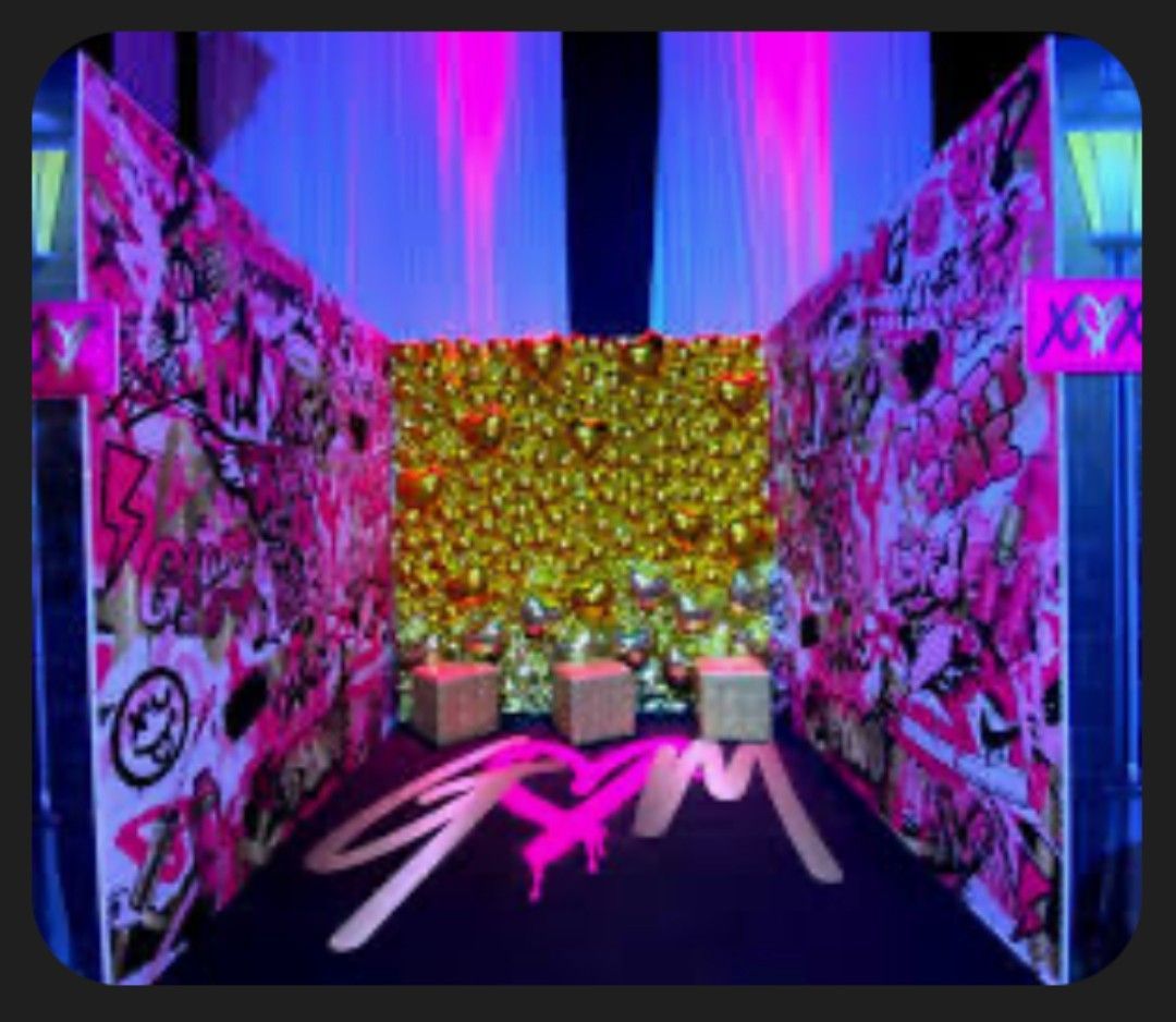 Pink and black graffiti-covered walls frame a backdrop of gold balloons. Three cube seats in front.