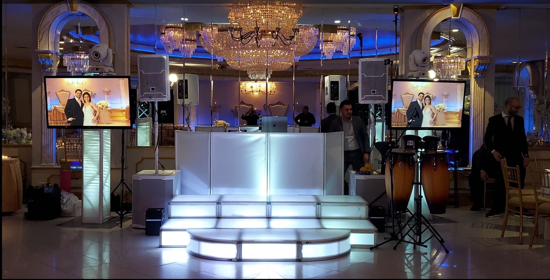 DJ setup at event. Stage with lights, screens showing photos, speakers, and a large chandelier.