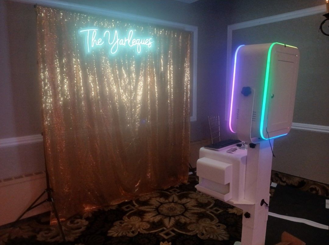 Photo booth with gold sequin backdrop and neon sign. Booth has colorful lights.