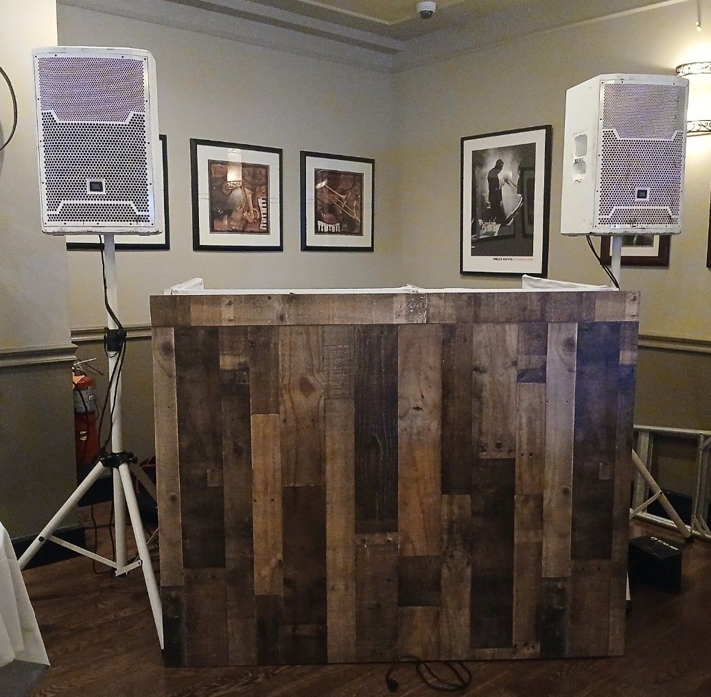 DJ booth with wooden paneling, two speakers on stands, and framed artwork on the wall.