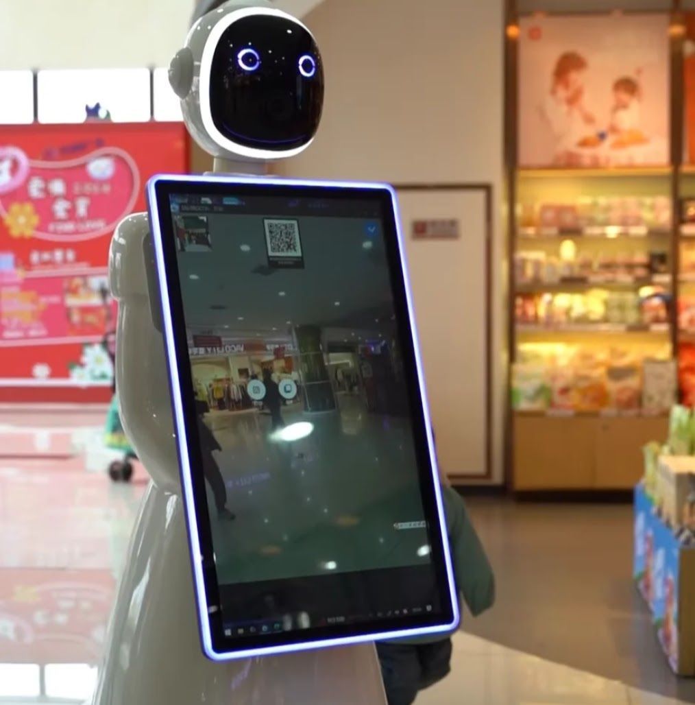 Robot holding a tablet displays a shopping mall scene. White body, black face with glowing eyes, standing indoors.