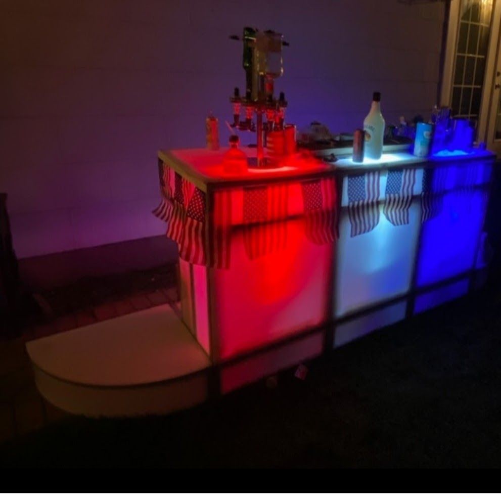 Lighted portable bar, red, white, and blue, set up outdoors at night with bottles and bar accessories.