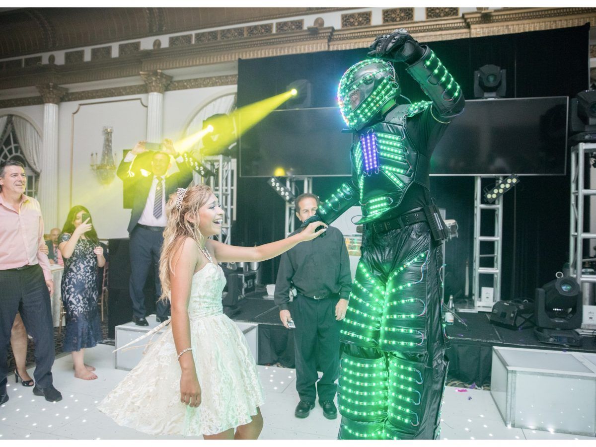A girl dances with a lit-up robot at a party; guests and stage lights are visible.