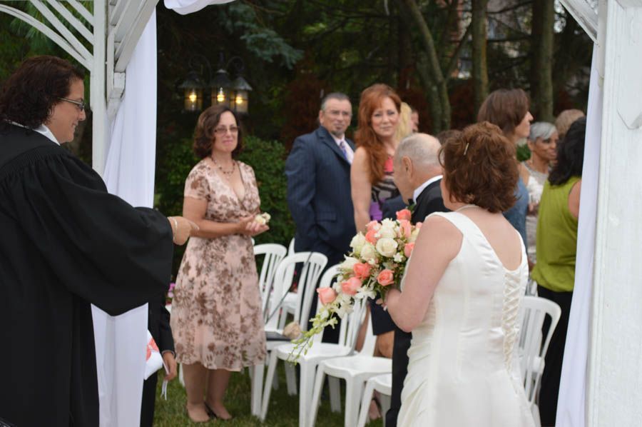 Wedding ceremony: Bride and groom, with officiant, guests, and decorated archway in garden setting.