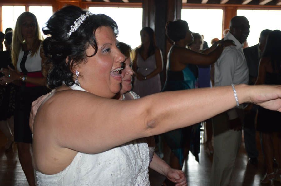 Bride dances with arm outstretched, smiling. Others in background dance in a room with large windows.
