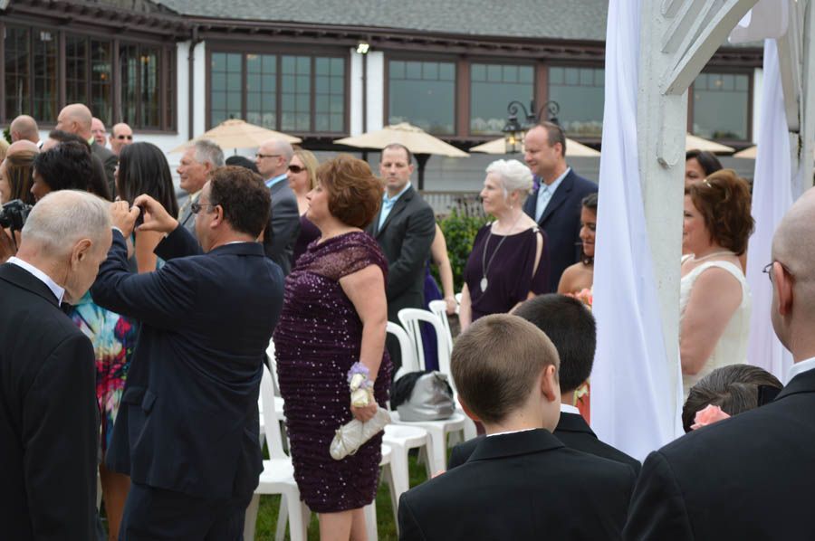 Wedding ceremony with guests, partially outdoors. People stand in front of a white trellis structure.