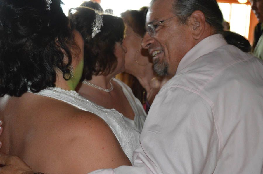Man smiles while hugging two women in white dresses; outdoors, wedding.