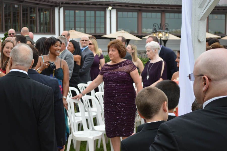 People gathered outdoors for a ceremony. Woman in purple dress directs guests to white chairs. Others watch.