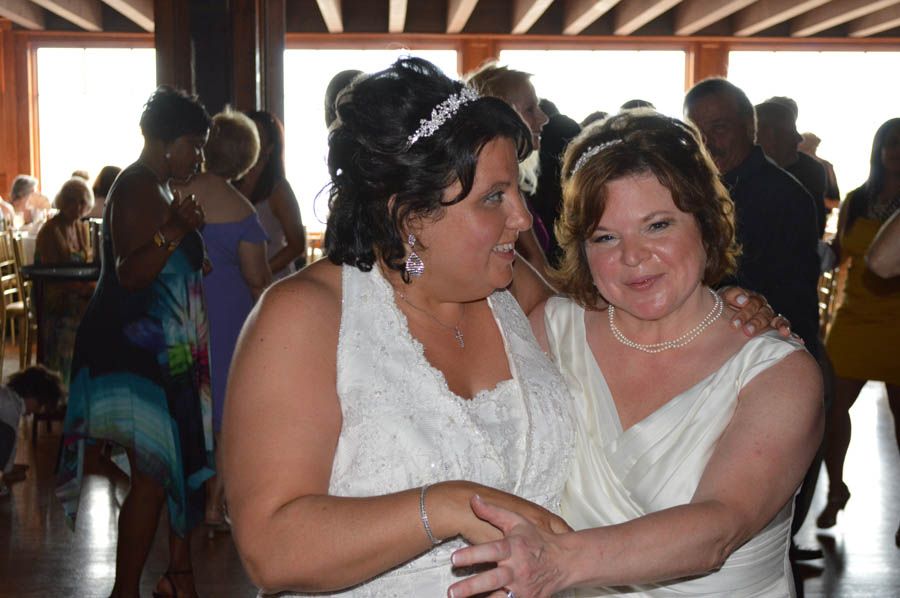 Two women in white dresses embrace indoors, surrounded by wedding guests.