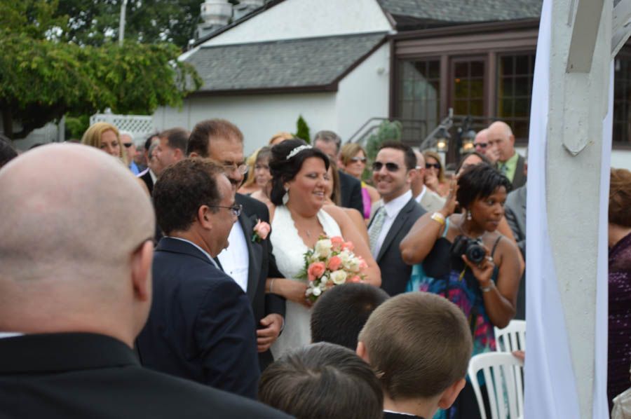 Bride walking down the aisle, smiling, with an escort. Outdoor wedding, guests watching.