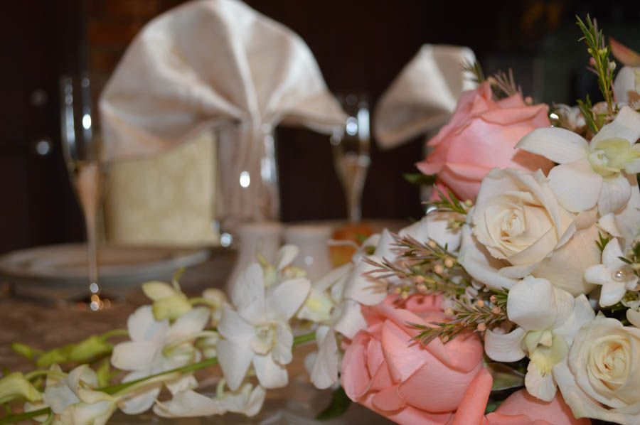 Close-up of pink and white floral arrangement on a table, with champagne flutes and folded napkin in the background.