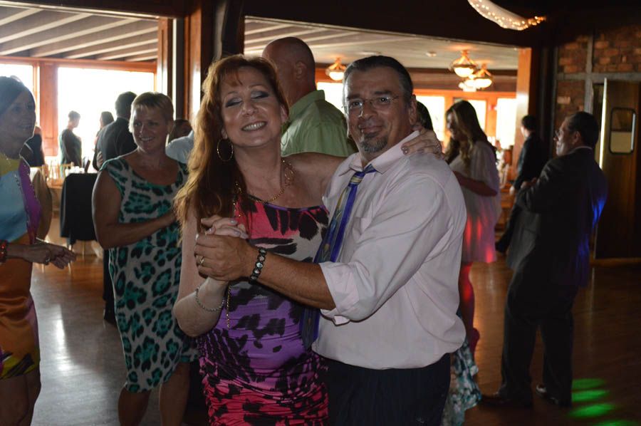 Couple dancing at a reception, smiling. Woman in floral dress, man in a pink shirt, others in background.