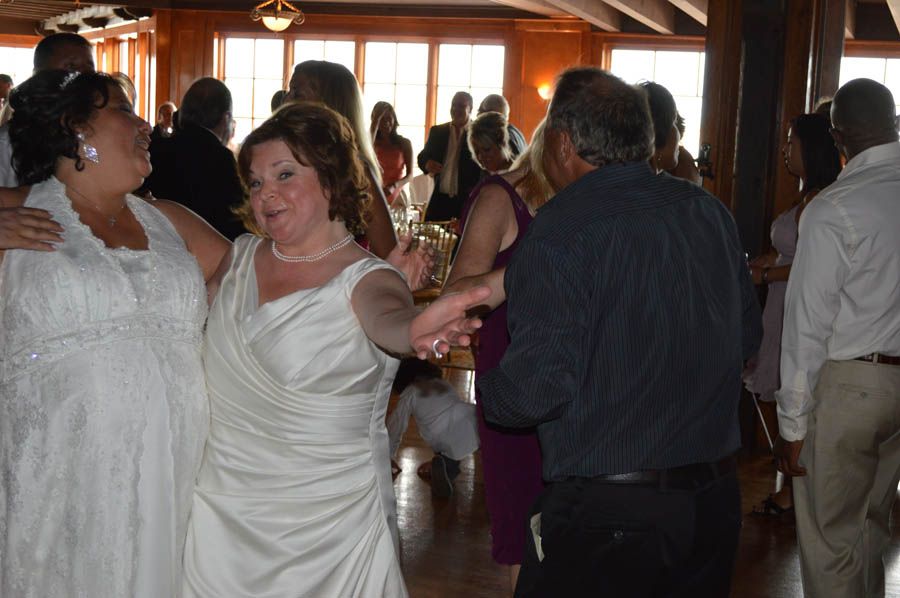 Two women in white dresses dance with a man in a formal setting, possibly a wedding reception.