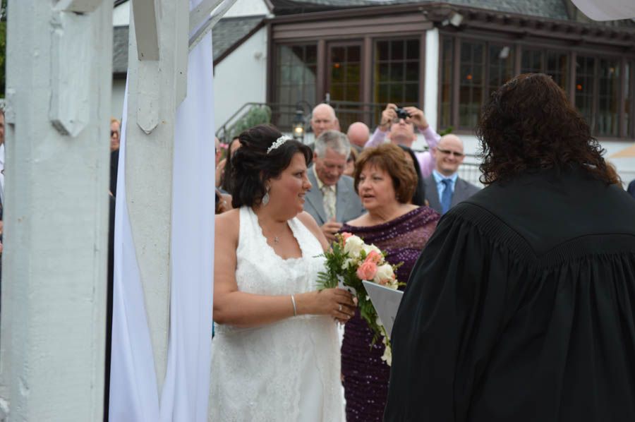 Bride and officiant at outdoor wedding ceremony; white structure, guests in background.