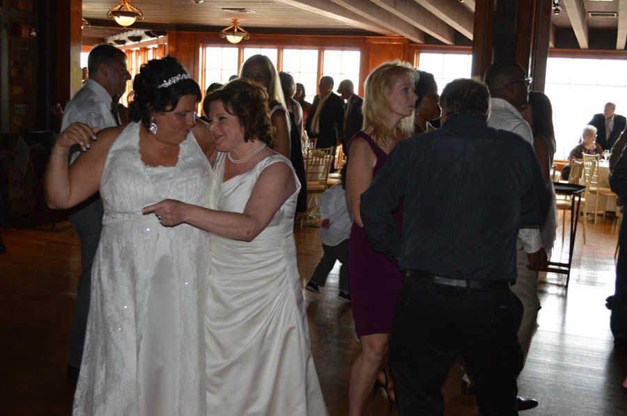Two women in wedding dresses dancing at a reception, others watch and dance nearby.