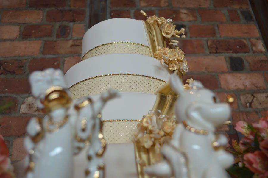 Three-tiered white and gold wedding cake with flower decorations; ceramic figurines in foreground.