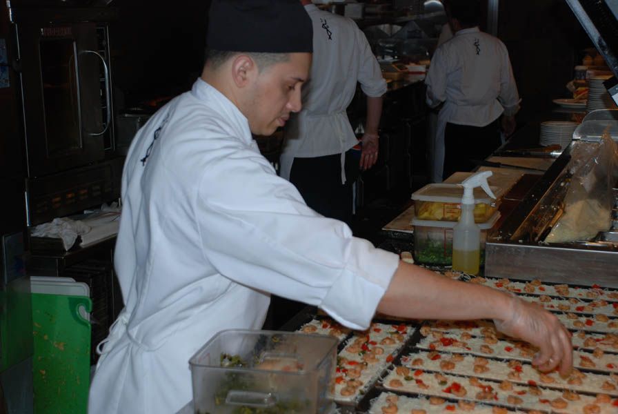 Chef in white coat and black hat placing toppings on flatbreads in a restaurant kitchen.