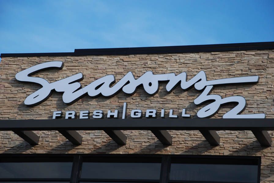 Seasons 52 Fresh Grill sign on a stone building, with blue sky above.