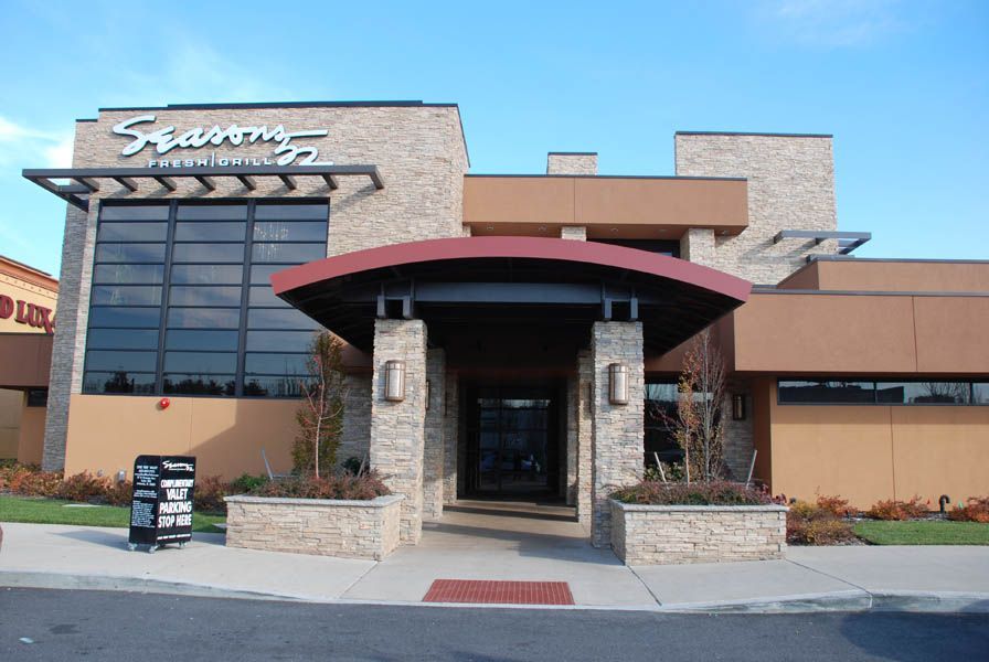 Seasons 52 restaurant exterior: tan and brown facade, entrance with red awning, large windows, blue sky.