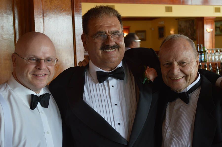 Three men in tuxedos, smiling at the camera indoors, possibly at an event.