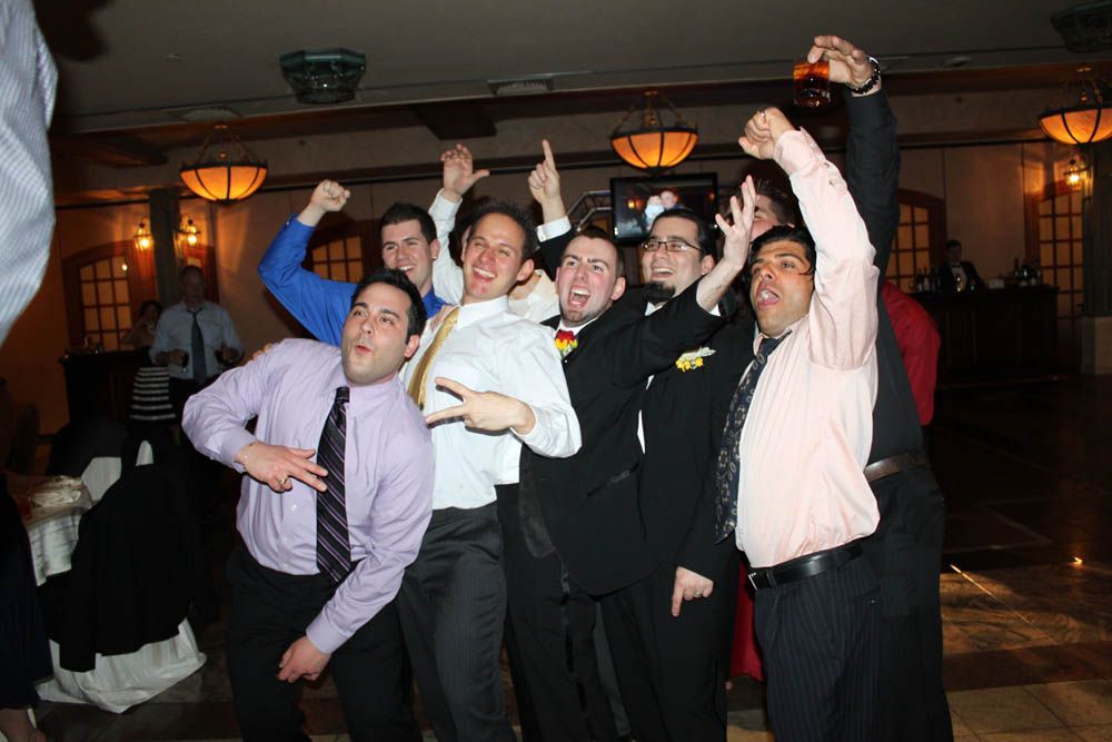 Group of men celebrating, arms raised, in a ballroom. They wear suits and dress shirts, joyful expressions.