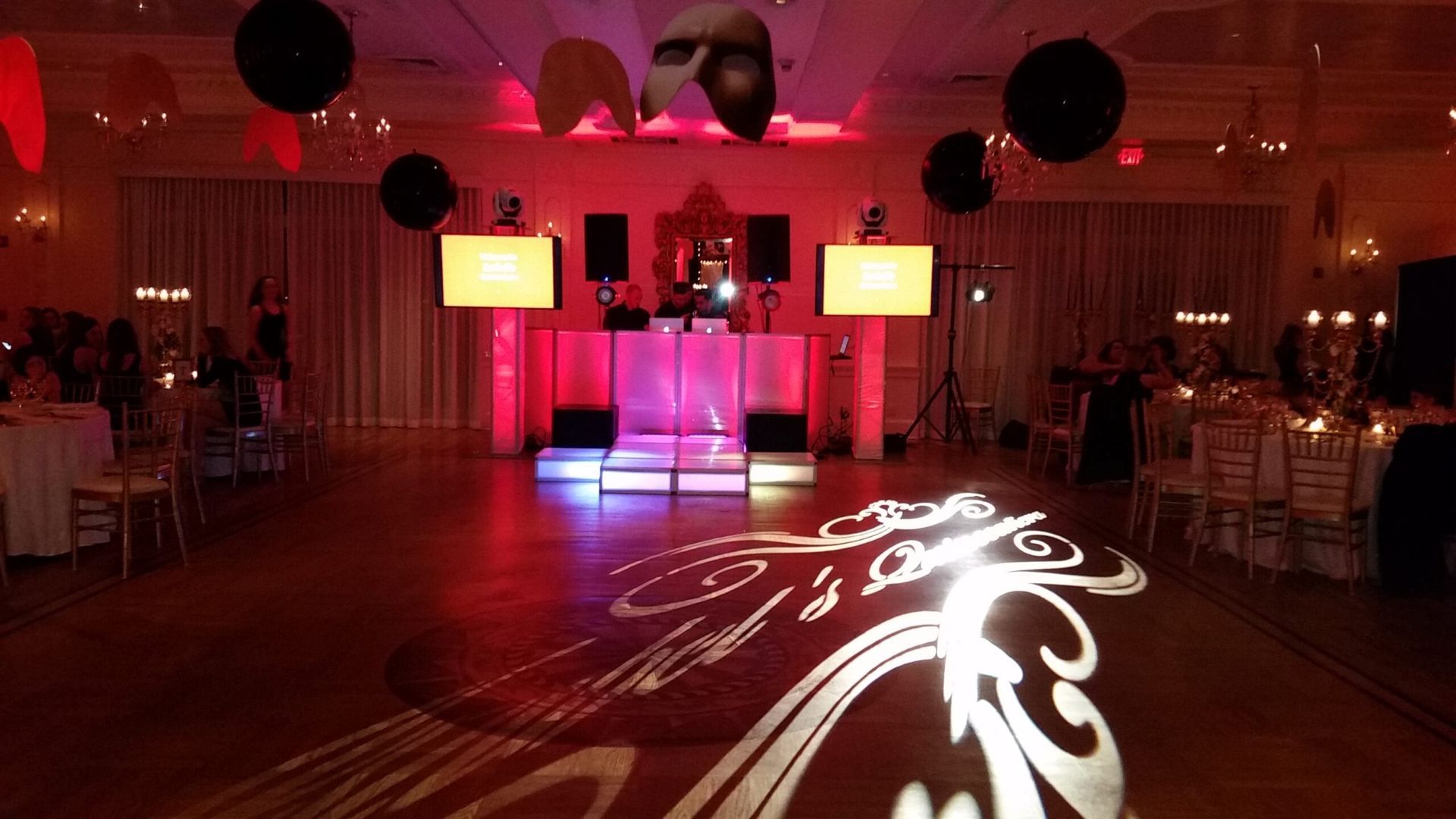 A ballroom with a DJ setup, dance floor projection, and tables set for a party, lit in red.