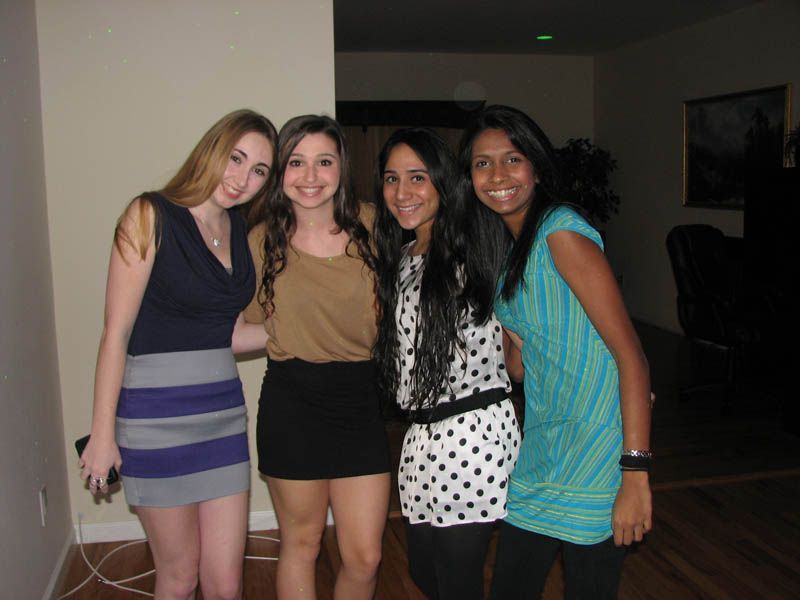 Four young people pose indoors, smiling. They wear party attire, hugging near a wall.