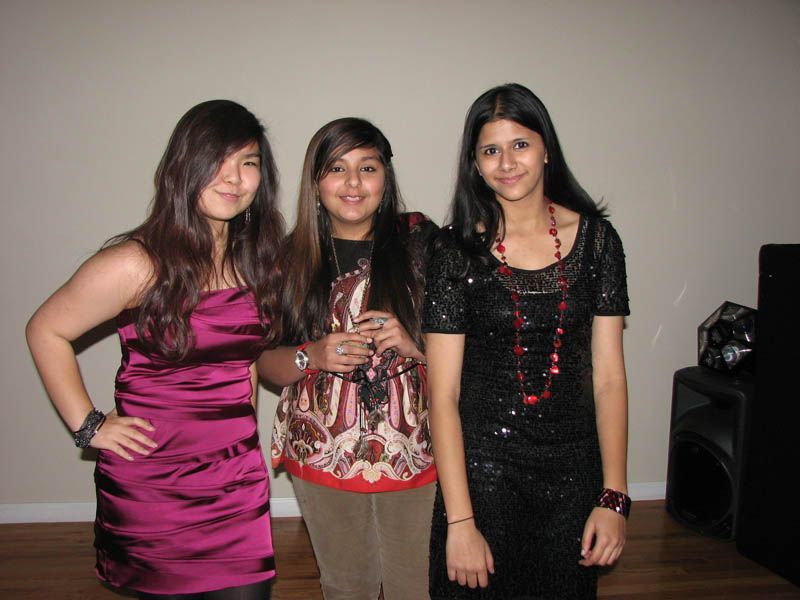 Three young women posing indoors. One wears a pink dress, another a patterned top, and the last a black sequined dress.