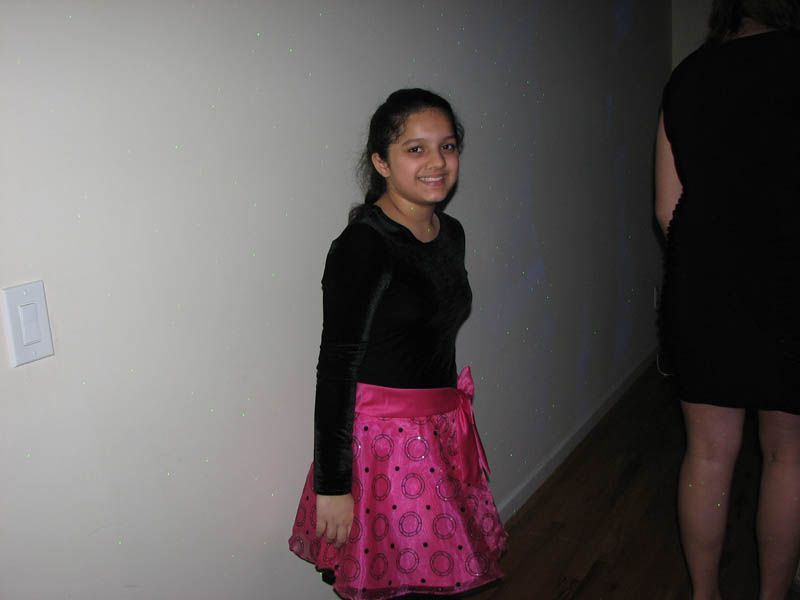 Woman in black top and pink skirt smiles near a wall. Another person in a black dress is partially visible.