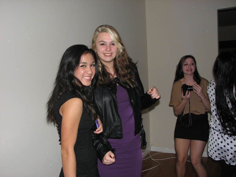 Three smiling women pose at a party. Two are in the foreground, and one holds a camera in the background.