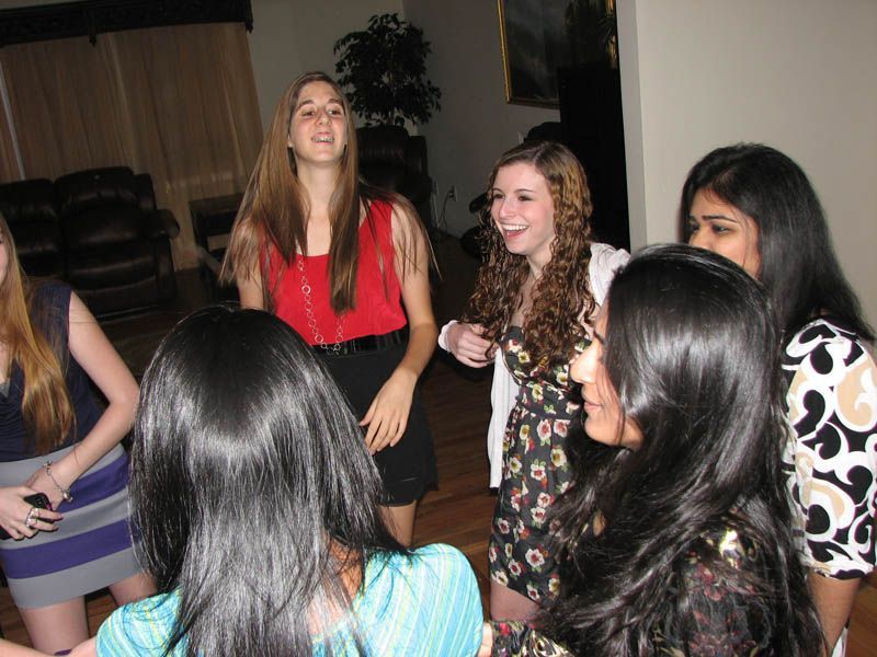 Group of young people laughing, standing in a circle indoors. Some wear dresses, appear to be at a gathering.