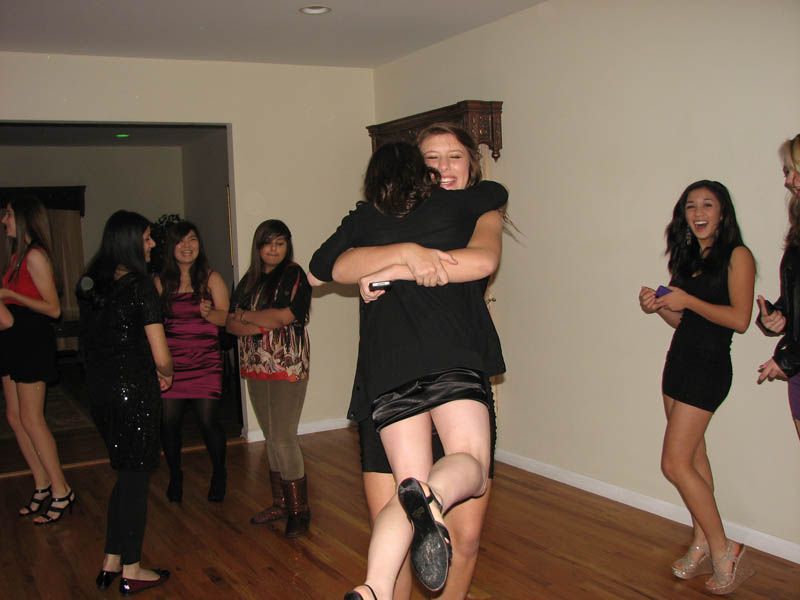 Two women embracing at a party; other women watch in a well-lit room.