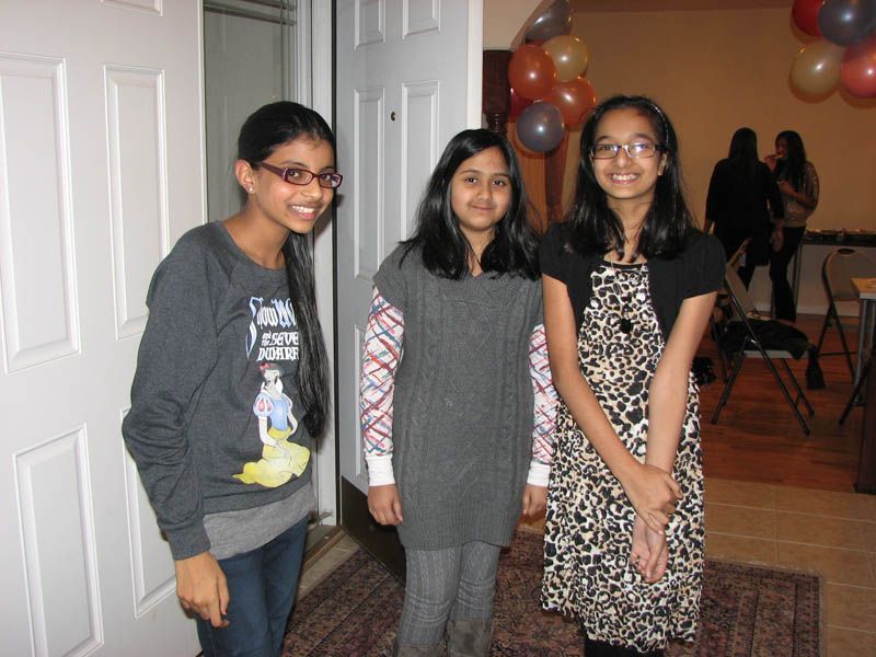 Three girls smiling near a doorway; one in a patterned dress, others in sweaters; balloons in the background.