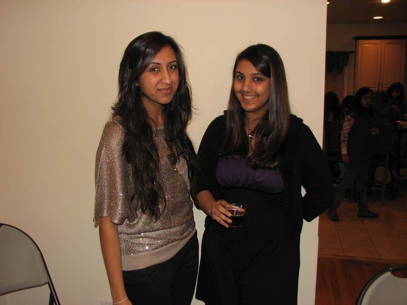 Two women stand indoors, one in a sequined top and the other in a black cardigan, smiling.