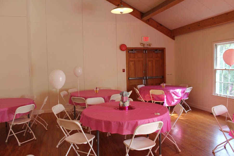 Pink-covered tables with white chairs in a room with balloons, set for a gathering.