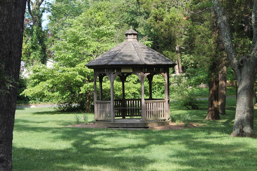 Wooden gazebo in a grassy park, surrounded by trees with lush green foliage.