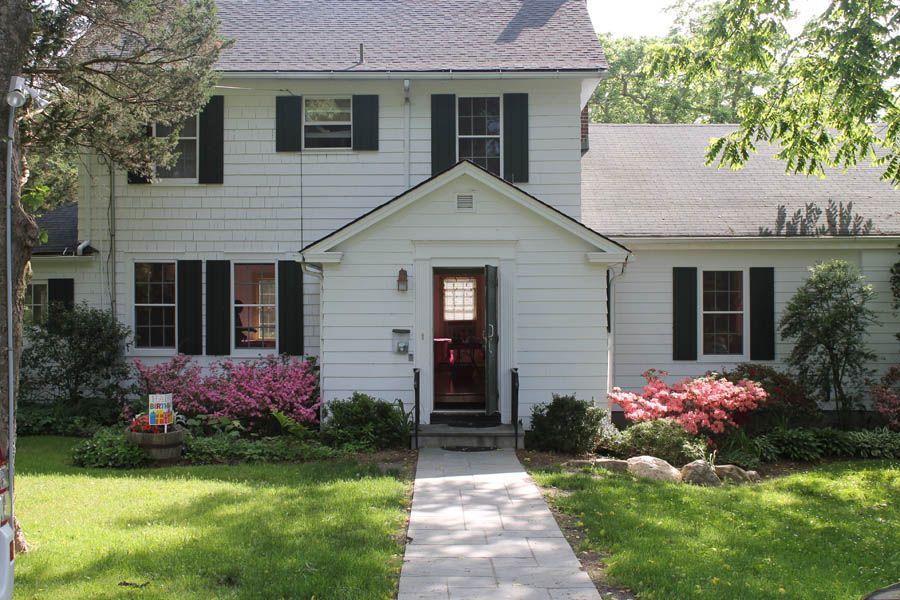 White two-story house with black shutters, a small porch, and a walkway. Pink flowers and green lawn.