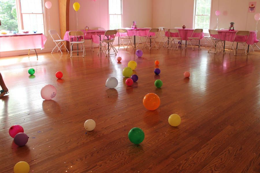 Balloons scattered on a wooden floor in a pink-decorated party room with tables and chairs.