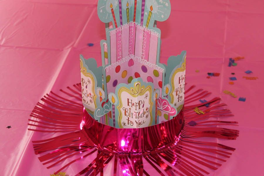Pink birthday cake centerpiece on a pink tablecloth with confetti.
