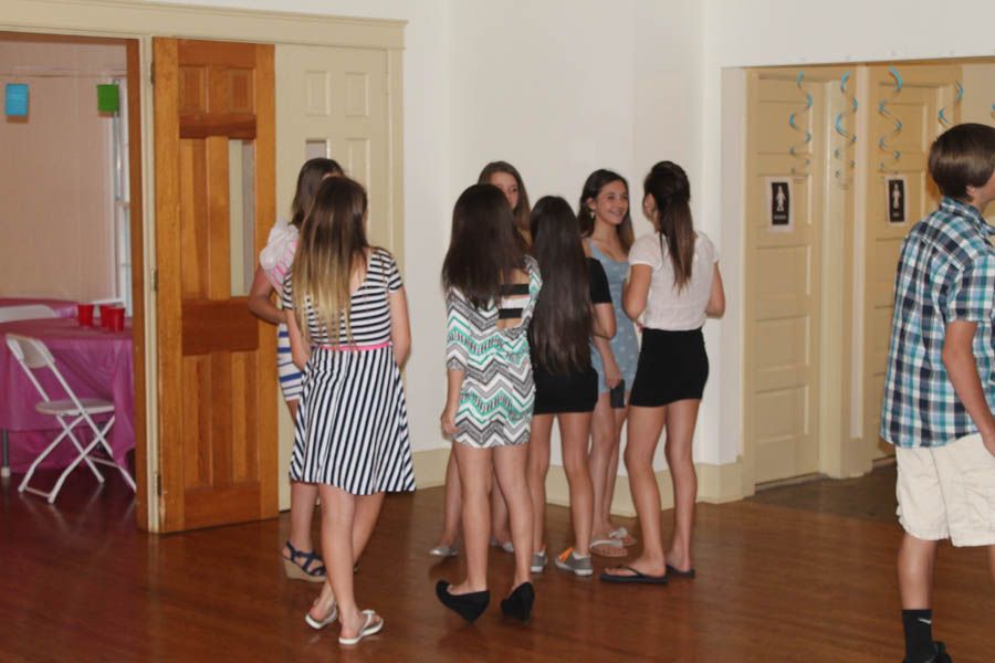 A group of young people gather inside a room with wooden floors. Some are wearing dresses and shorts.