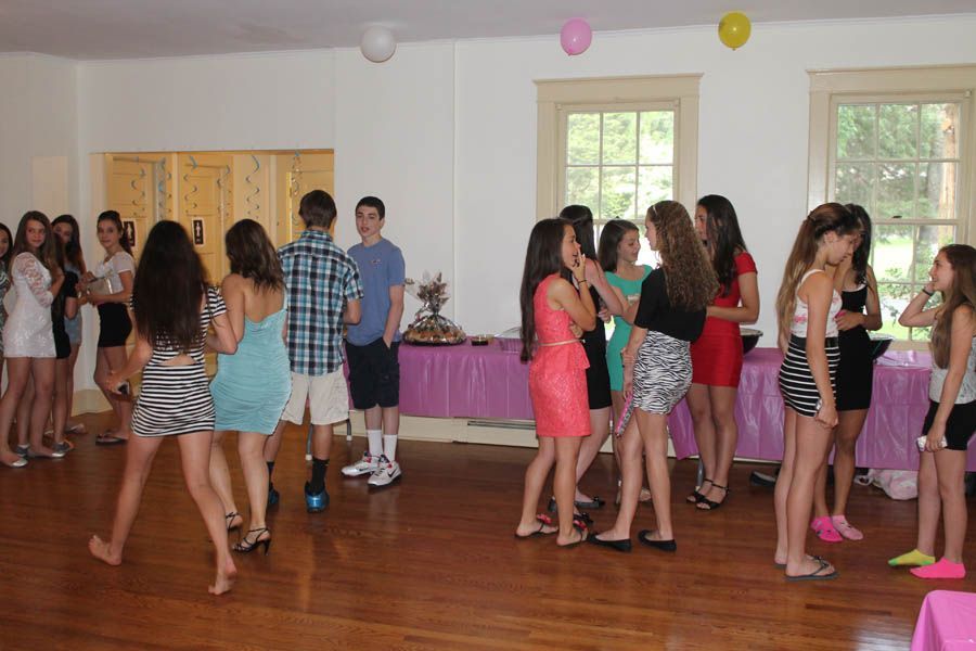 A group of teens in party attire mingle in a room decorated with balloons and a buffet table.