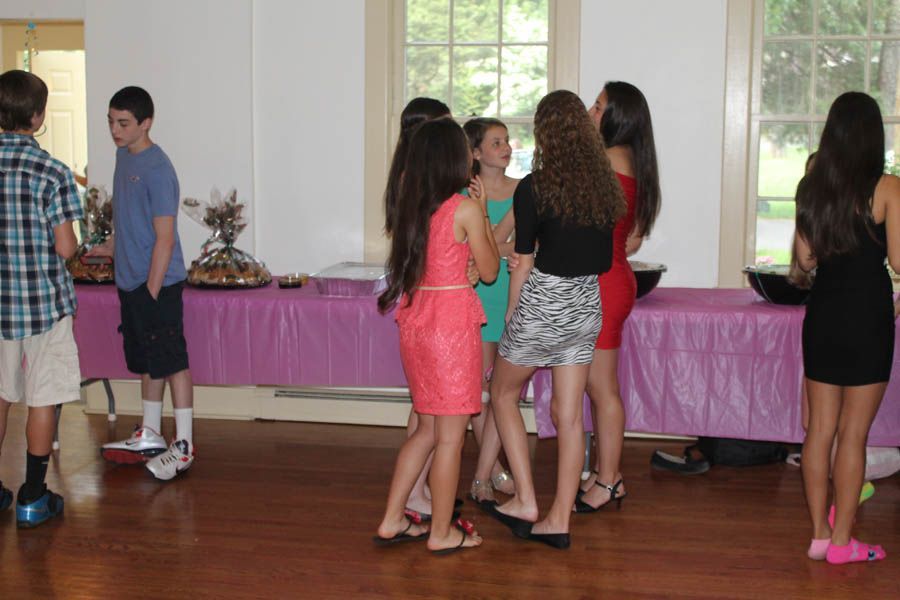Teenagers gathered at a party near a table with food, in a room with windows and hardwood floors.