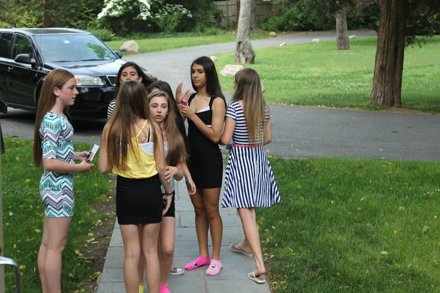 Six young people stand near a driveway. A dark SUV is parked nearby. Girls in summer outfits are talking.