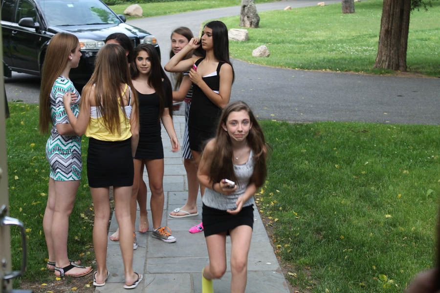 Group of girls on a sidewalk near a driveway. One girl looks toward the camera while holding a phone.