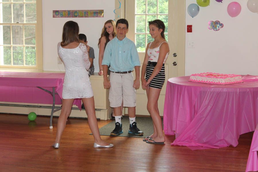 Teenagers at a party: girl in white dress poses, boy in shorts and others stand near pink tables, balloons.