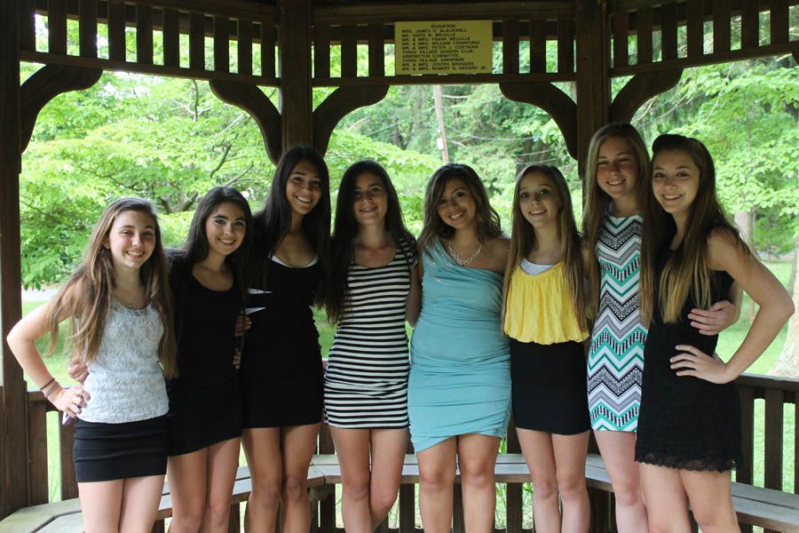 Group of young people smiling, standing closely together in a wooden gazebo, wearing various dresses.